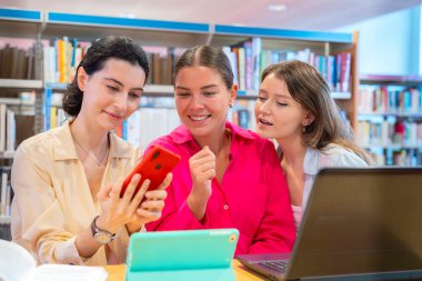Üç kadın kütüphanede birlikte oturur, gülümser ve bir telefon ve tablete bakarlar. Arka planda kitap rafları ve bir dizüstü bilgisayar görülebiliyor, canlı ve işbirlikçi bir çalışma ortamı yaratıyor.