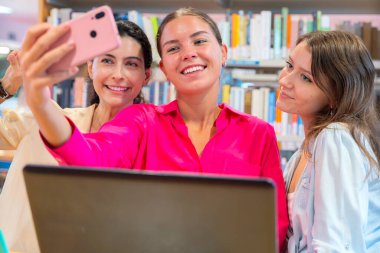 Üç kadın kütüphanede bir selfie için poz veriyor, gülümsüyor ve barış işaretleri gösteriyor. Kitap rafları arka planda, canlı ve eğlenceli bir atmosfer yaratıyorlar..