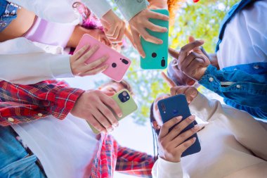  Grup A 'nın bir grup arkadaşı dışarıda ayakta duruyor, her biri akıllı telefon tutuyor. Telefonları farklı renklerde, bağlantıları ve takım çalışmalarını gösteriyor. Açık havada.