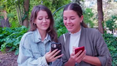 İki arkadaş dışarıda akıllı telefonlarını kullanırken birlikte vakit geçirmekten zevk alıyor. Bereketli yeşilliklerle çevrili olarak, rahatlatıcı bir park ortamında günlük ve neşeli bir etkileşimi paylaşıyorlar..