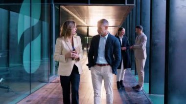 İki iş adamı sohbet ederken cam duvarlı bir ofis koridorunda yürürler. Arka planda, meslektaşlar etkileşime geçerek, bir işbirliği ve verimlilik ortamı yaratıyorlar..