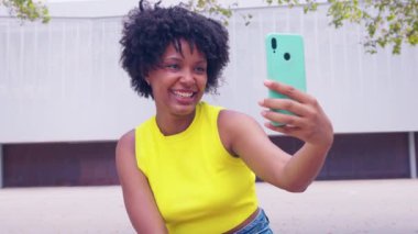 Açık havada alışveriş yaparken gülümseyen ve akıllı telefonuyla selfie çeken Afrikalı Amerikalı bir kadın. Renkli alışveriş torbaları taşıyorlar ve şehir ortamında güneşli bir günün tadını çıkarıyorlar.