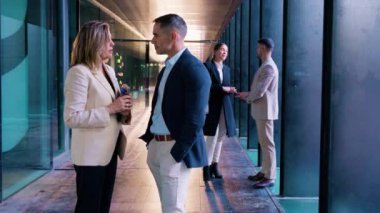 İki iş adamı sohbet ederken cam duvarlı bir ofis koridorunda yürürler. Arka planda, meslektaşlar etkileşime geçerek, bir işbirliği ve verimlilik ortamı yaratıyorlar..