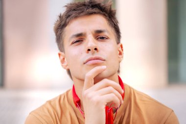 Kendinden emin ve düşünceli bakışları olan genç bir adam çenesini elinin üzerine koyuyor. Boynuna kırmızı kulaklık takıyor ve sıradan bir tişört giyiyor. Açık hava kentsel ortamında kameraya bakıyor..