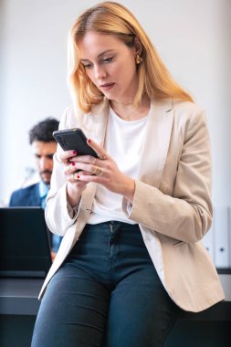  Professional woman in a suit concentrating on his smartphone in a bright office. A female colleague works in the background, creating a collaborative work environment. clipart