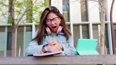  Gözlük takan ve pembe kulaklık takan genç bir kadın açık havada defter ve tabletle çalışırken esniyor. Arka planda ağaçlar ve binalar olan modern eğitim ortamı.
