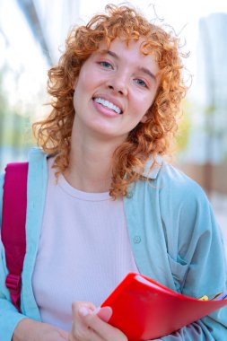 Neşeli, genç, kızıl saçlı, günlük kıyafetler giyen, sırt çantalı bir kadın kitap ve akıllı telefon tutuyor. Parlak bir şehir ortamında, kendine güveni ve pozitifliği yayarak dışarıda duruyor..
