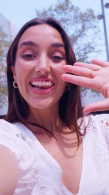 Uzun kahverengi saçlı neşeli genç bir kadın açık havada selfie videosu çekerken kameraya merhaba diyor. Beyaz bir bluz giymişti. Arka planda ağaçlarla birlikte gün ışığında duruyordu..