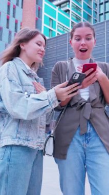 Modern bir şehir ortamında dışarıda duran iki genç kadın akıllı telefonlarına bakarken heyecanla gülümsüyor ve tepki veriyorlar. Biri kot ceket giyiyor, diğeri şık gri ceket..