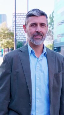 Neşeli genç bir adamın portresi, yüksek binalar ve canlı mimari ile modern bir şehir ortamında güvenle gülümsüyor..