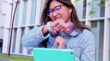  Boynunda gözlük ve kulaklık olan genç bir kadın dışarıda otururken konuşmak için akıllı kasığını kullanıyor. O, modern bir ortamda yanında bir tablet ile sıradan bir kot ceket giydi..