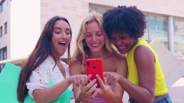 Üç neşeli genç kadın kırmızı bir akıllı telefonun etrafında toplanıyor, heyecanla tepki veriyorlar. Gelişigüzel giyiniyorlar, modern bir şehir ortamında dışarıda duruyorlar, birlikte eğlenceli bir anın tadını çıkarıyorlar..