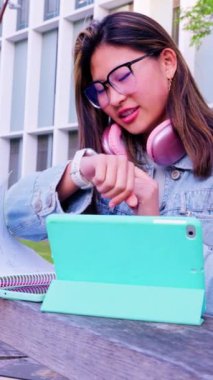  Boynunda gözlük ve kulaklık olan genç bir kadın dışarıda otururken konuşmak için akıllı kasığını kullanıyor. O, modern bir ortamda yanında bir tablet ile sıradan bir kot ceket giydi..