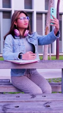 Pembe kulaklık takan ve kot ceketli şık bir kadın merdivenlerde selfie için poz veriyor. Neşeli ifadesi ve günlük kıyafetleri modern yaşam tarzını, özgüvenini ve açık havada kendini ifade edişini vurguluyor.