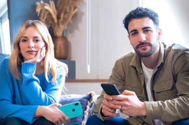 Akıllı telefonlar kullanarak evde oturan genç bir çift düşünceli duygularını ifade ediyorlar. Kadın telefonunu düşünceli bir bakışla tutarken, erkek tarafsız bir ifadeyle bakıyor.