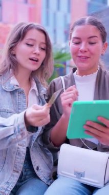İki şık genç bayan dışarıda oturmuş, internetten alışveriş yapıyorlar. Biri kredi kartı taşırken diğeri yeşil tablet kullanıyor. Canlı bir şehir ortamında heyecan verici bir anı paylaşıyorlar..