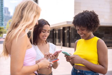 Üç neşeli genç kadın kırmızı bir akıllı telefonun etrafında toplanıyor, heyecanla tepki veriyorlar. Gelişigüzel giyiniyorlar, modern bir şehir ortamında dışarıda duruyorlar, birlikte eğlenceli bir anın tadını çıkarıyorlar..