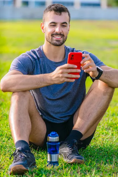 Genç atletik bir adam parkta yeşil çimlerde bağdaş kurmuş, elinde kırmızı bir akıllı telefon tutuyor. Rahatlamış görünüyor, gülümsüyor ve yanında bir su şişesi ile spor giysiler giyiyor. Dikey
