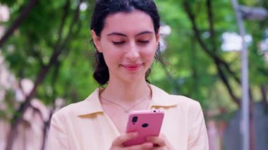 Gülümseyen genç bir kadın pembe akıllı telefonunu kullanırken rahat bir şekilde bankta oturur. Günlük kıyafetlerle mutlu ve rahat görünüyor. Etrafı tuğla duvarlarla çevrili..
