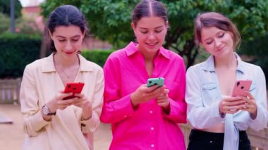 Açık havada birlikte oturan üç genç kadın, yeşil ve çiçeklerle çevrili akıllı telefonlarıyla gülümsüyor ve içeriklerini paylaşıyorlar..