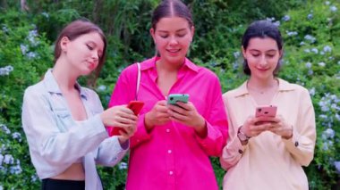 Açık havada birlikte oturan üç genç kadın, yeşil ve çiçeklerle çevrili akıllı telefonlarıyla gülümsüyor ve içeriklerini paylaşıyorlar..