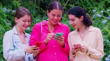Açık havada birlikte oturan üç genç kadın, yeşil ve çiçeklerle çevrili akıllı telefonlarıyla gülümsüyor ve içeriklerini paylaşıyorlar..