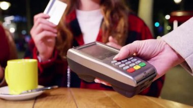 Kadınlar geceleri açık bir kafede oturuyor, temassız bir şekilde kredi kartıyla ödeme yaparken gülümsüyorlar. Kahveler ahşap masada..