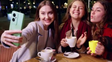 Üç gülümseyen kadın gece bir kafede selfie çekiyor, ellerinde kahve fincanları ve mutlu poz veriyorlar. Dışarıda çekilen eğlenceli ve neşeli bir an..