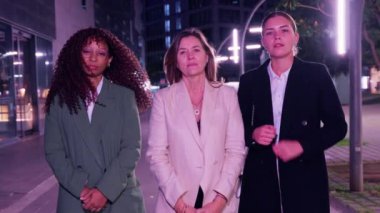 Üç kendine güvenen iş kadını gece dışarıda kolları bağlı, ciddi bir şekilde dikiliyorlar. Profesyonel kıyafetler giyiyorlar. Arka planda modern şehir ışıkları ve ofis binaları var..