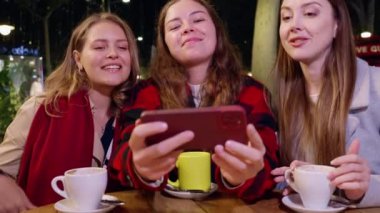 Açık bir kafede otururken gülümseyen ve selfie çeken üç genç kadın. Kahvenin tadını çıkarıyorlar ve sıcak bir atmosferde keyifli bir anı paylaşıyorlar.