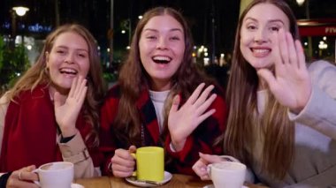Üç neşeli kadın gece açık bir kafede oturuyor, kameraya el sallıyor ve gülümsüyor, kahve fincanları ahşap masada. Sıcak ve dostane bir atmosfer..