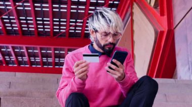 Saçları boyalı ve gözlüklü genç bir adam elinde kredi kartı ve akıllı bir telefonla merdivenlerde oturuyor, online alışveriş ya da ödeme yapıyor. Kırmızı mimari arka plana sahip modern kentsel ortam.