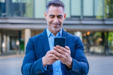 Smiling middle-aged businessman in a blue suit holding and using a smartphone outdoors in a modern urban setting. clipart