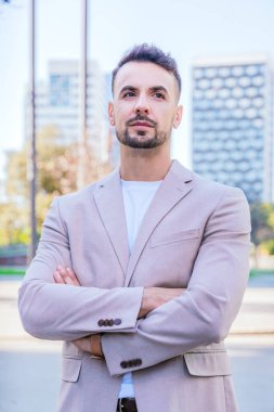 Portrait of a young businessman in a beige blazer standing outdoors in an urban setting with arms crossed, exuding confidence and professionalism clipart