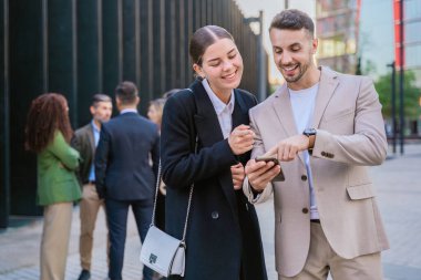 Akıllı telefona bakarken gülümseyen iki genç iş arkadaşı. Arka planda diğer profesyonellerle birlikte modern bir şehir ortamında dışarıda duruyorlar..