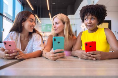 Modern bir kafede masada oturan, ellerinde akıllı telefonlar olan ve gülümseyen üç farklı kadından oluşan bir grup. Arkadaşlık ve dijital bağlantıyı temsil eden rahat ve neşeli bir atmosfer..