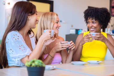 Üç farklı kadından oluşan bir grup bir masada oturuyor, ellerinde kahve fincanları, ve sıcak bir kafede birlikte gülüyorlar. Sıcak ve neşeli atmosfer, dostluğu ve bağlantıyı simgeliyor..