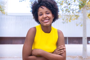 A cheerful young woman with natural curly hair wearing a yellow top smiles confidently while posing outdoors. Bright and vibrant setting, showcasing joy, beauty, and individuality. clipart