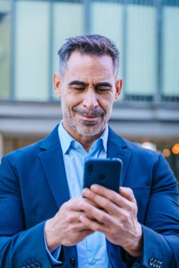 Smiling middle-aged businessman in a blue suit holding and using a smartphone outdoors in a modern urban setting. clipart