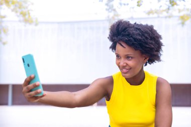  Sarı bluzlu, kıvırcık saçlı neşeli genç bir kadın açık havada selfie çekiyor, gülümsüyor ve barış işareti yapıyor. Canlı sahne mutluluk, bireysellik ve modern yaşam tarzını gösteriyor. Kopya alanı