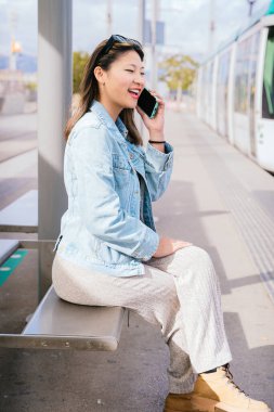 Sıradan kıyafetler ve kot ceket giyen gülümseyen genç bir kadın tramvay durağında oturuyor ve cep telefonuyla konuşuyor. Tramvay ve şehir geçmişi olan şehir merkezi. Parlak ve güneşli bir gün.