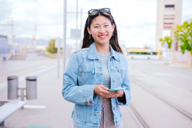 Kot ceketli neşeli genç bir kadın mutlu bir ifadeyle yukarı bakarken akıllı telefonunu tutuyor. Modern kentsel ortam, dijital bağlantıyı ve şehir yaşam tarzını temsil ediyor.