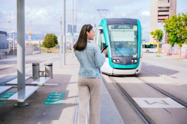 Tramvay durağında duran kot ceketli genç bir kadın yaklaşan tramvayı beklerken akıllı telefonunu tutuyor. Bir şehir ortamında modern şehir ulaşımı ve gidiş-dönüş kavramı.