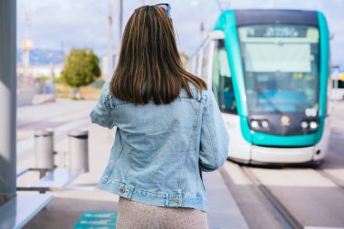 Tramvay durağında duran kot ceketli bir kadın yaklaşan bir tramvaya bakıyor. Modern bir şehir ortamında toplu taşıma için bekliyor. Yolculuk ya da yolculuk için hazır..
