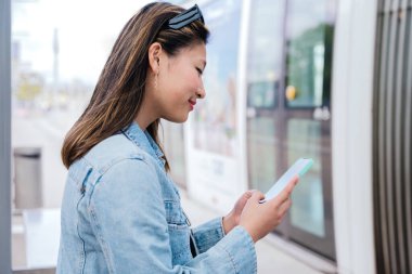 Tramvaya binmek için beklerken akıllı telefonunu kullanan kot ceketli genç bir kadının yan görüntüsü. Modern şehir ulaşımı ve dijital bağlantı kavramı.