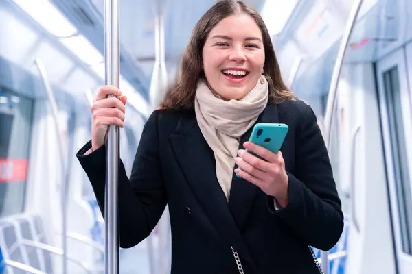 Siyah paltolu ve bej eşarplı genç bir kadın modern bir metro treninin içinde elinde akıllı bir telefonla metal bir direği kavrıyor. İşe gidip gelirken odaklanmış görünüyor. Elinde şık beyaz bir çanta var.. 