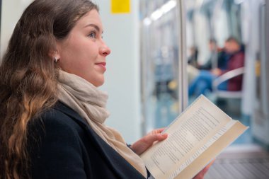 Siyah paltolu ve bej eşarplı genç bir kadın modern bir metro treninde oturmuş, odaklanmış bir kitap okuyor. Şehirdeki seyahati sırasında sakin bir anın tadını çıkarıyor.. 