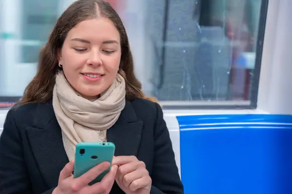 Siyah ceketli ve bej eşarplı neşeli genç bir kadın modern bir metro treninde oturmuş akıllı telefonuna bakıp gülüyor. Seyahat sırasında eğlenceli bir anın tadını çıkarıyor.. 