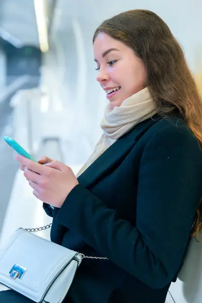  Modern bir metro istasyonunda oturan siyah ceketli ve atkılı şık genç bir kadın akıllı telefonunu kullanırken gülümsüyor. Parlak ve temiz ortam şehir ortamını güçlendirir. 