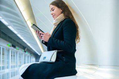  Modern bir metro istasyonunda oturan siyah ceketli ve atkılı şık genç bir kadın akıllı telefonunu kullanırken gülümsüyor. Parlak ve temiz ortam şehir ortamını güçlendirir. 